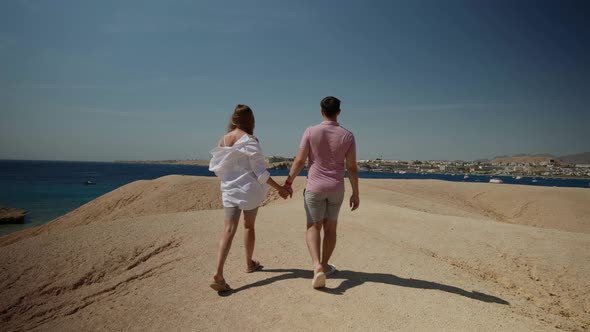 a Man in a Pink Tshirt and a Woman with Long Hair in a White Shirt Walk Away Along a Deserted Sandy alt