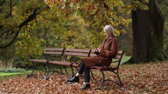 Woman with mobile phone siting on a bench in autumn park