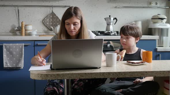 Mother Telecommuting on Laptop Next to Her Little Son Having Breakfast in the Kitchen