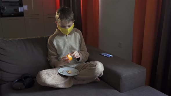 A Teenage Guy in a Protective Mask Peels a Tangerine While Sitting on a Sofa. Coronavirus Epidemic