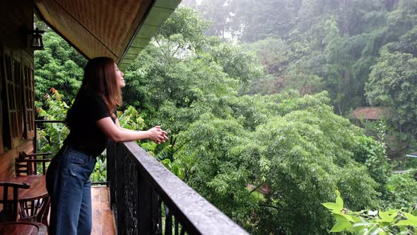 A woman standing on balcony, looking and touching the rain