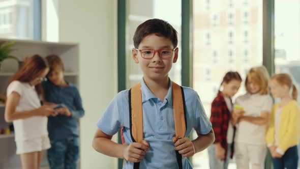 Portrait of the Smiled Asian Pupil Wearing Glasses and Carrying Backpack Standing in the Class While alt