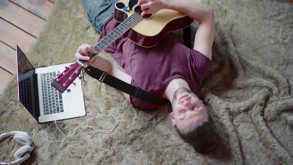 Man with Acoustic Guitar Singing and Playing Guitar Lying Floor Near Laptop and Headphones alt