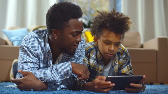 African American Man and Son at Home Lying on Floor in Living Room and Using Tablet Pc alt