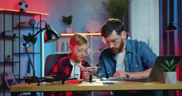 Dad in Protective Glasses Explaining How to Attach Details Using Soldering Iron for His Son at Home alt