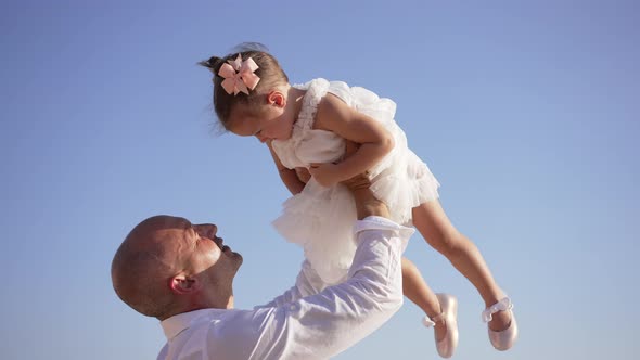 Side View of Confident Cheerful Father Raising Cute Little Girl in Dress at Background of Clear Blue alt