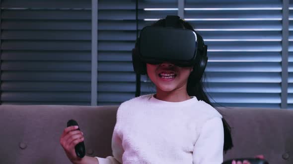Asian little girl having fun playing video game with virtual reality glasses on sofa at home. alt