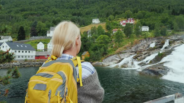 A Woman Admires the Norwegian Village and the Waterfall alt