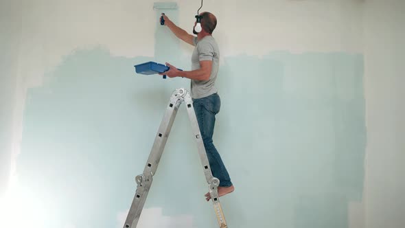 Caucasian Man Paints the Wall with a Roller Man Stands on a Stepladder Dad Makes Repairs in the Room