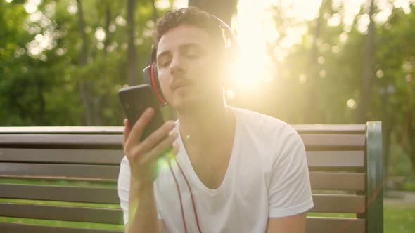 A Young Handsome Guy in a White Tshirt and Headphones Sits on a Bench in the Park at Sunset and alt