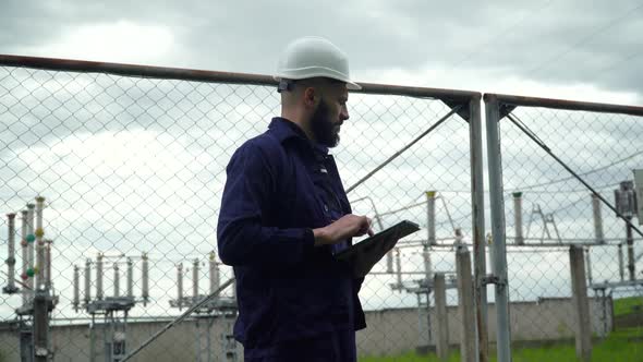 Engineer in Special Uniform Standing with Tablet Near Power Station. Male Monitoring System alt