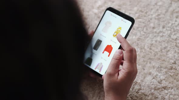 Woman Holding Smartphone Buying Fashion Clothes in Online Store at Cozy Home While Lying on Carpet alt