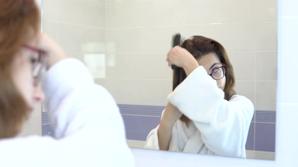 Young Woman with Glasses Combing Her Hair in Front of a Mirror. A Girl in a White Bathrobe in Front alt