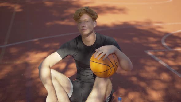 Exhausted Perspiring Redhead Sportsman Sitting with Ball on Outdoor Court and Looking Up at Sunlight alt