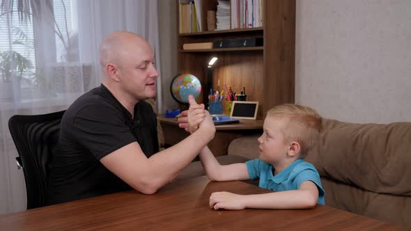 Happy Father is Engaged in Arm Wrestling with His Son at Home at the Table alt