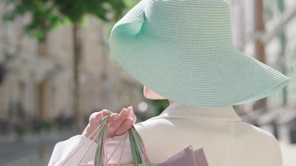 Close-up of Woman in Elegant Turquoise Hat Raising Shopping Bags on Shoulder and Turning To Camera alt