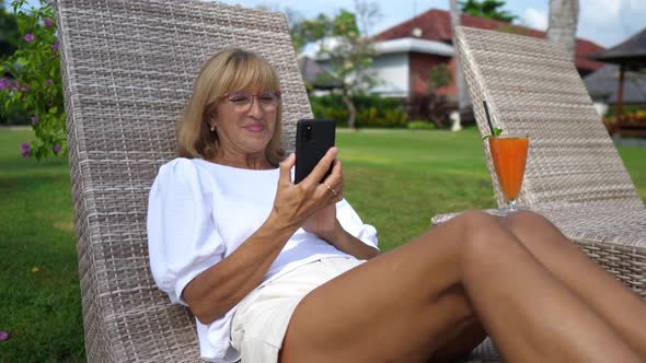 Woman Over 50 on a Sunbed Smiling at Her Smartphone While Video Calling with Beloved Ones alt