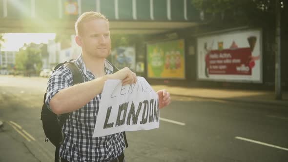 With a soft golden sunsetting glow surrounding him, a red-headed man holds up a sign hitchhiking to  alt