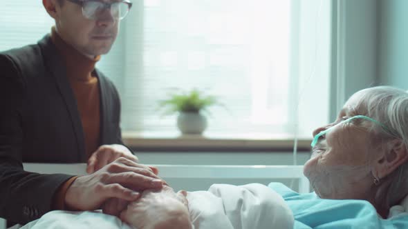 Son Supporting Elderly Mother in Hospital Ward alt