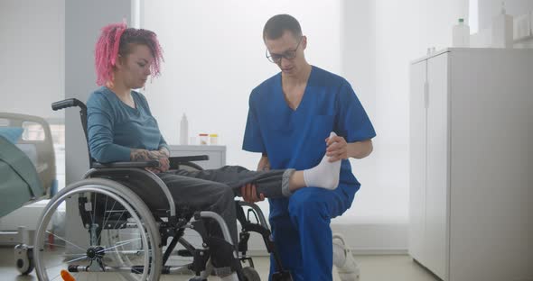 Doctor Holding and Massaging Young Woman Leg in Wheelchair at Fitness Center alt