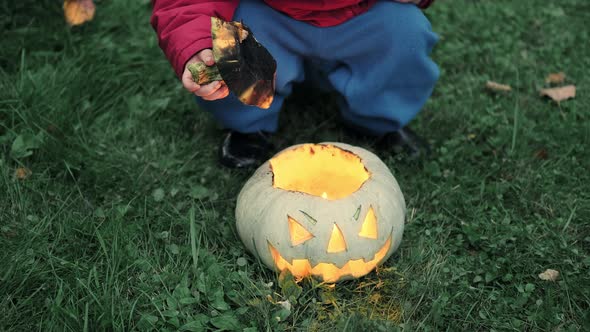 Little Girl in Jacket Plays with Glowing Pumpkin for Halloween on Grass in Park