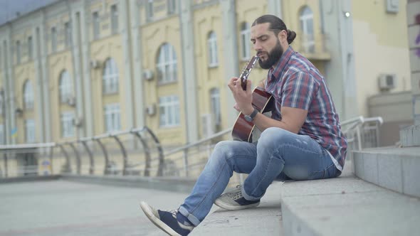 Wide Shot Side View of Young Caucasian Man Sitting on Urban Stairs and Playing Guitar. Handsome alt