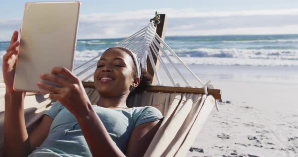 African american woman using digital tablet while lying on a hammock at the beach alt