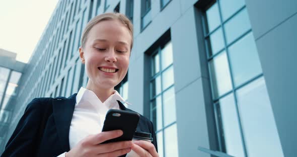Young Female is standing near Office Building, textng Messages on her Smartphone alt