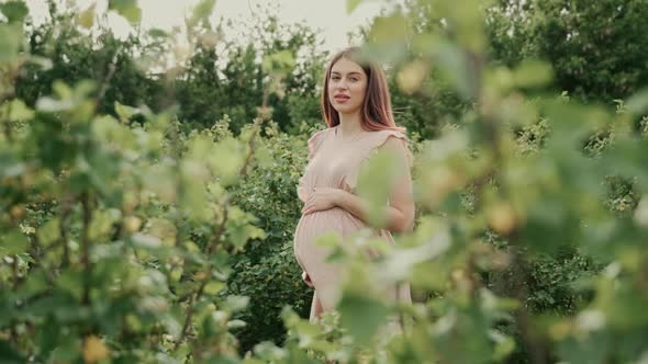 Young pregnant woman with flowing hair in a pink summer dress stands among the green plants. 