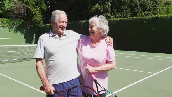Happy senior caucasian couple holding tennis rackets and embracing in tennis court alt