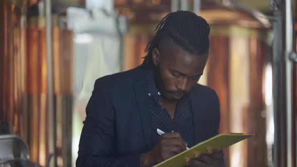 Expert African American Brewer Checking Beer Quality Filling in Paperwork Standing at Large Tanks alt