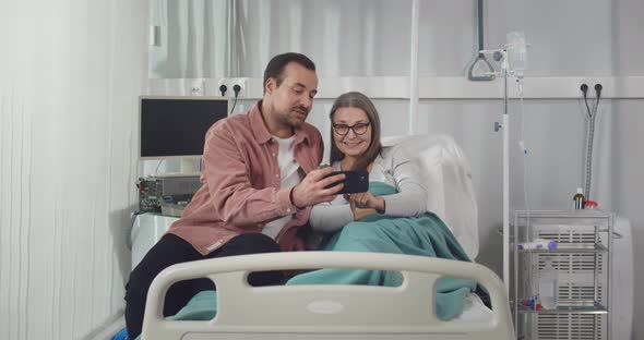 Young Man Visiting Senior Mother in Hospital Showing Family Photos on Smartphone alt