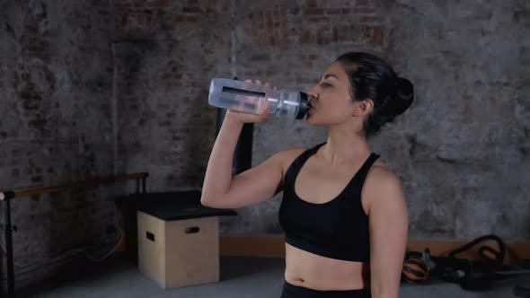 Athletic Indian girl drinks from a bottle water, woman in the gym enjoying fresh water alt