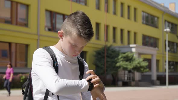 A Caucasian Teenage Boy Works on a Smartwatch  a School in the Background