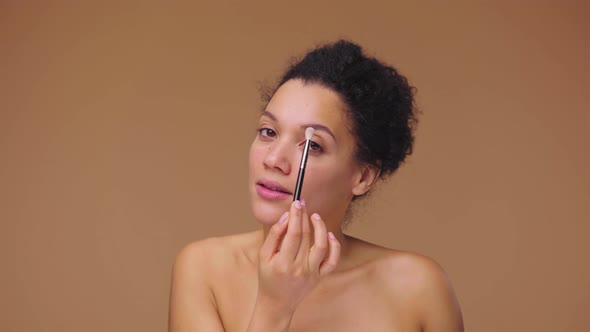 Beauty Portrait of Young African American Woman Looking in Mirror and Doing Eye Makeup Using Brush alt