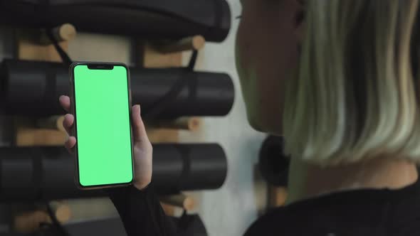 A Young Athletic Woman Holds A Smartphone in Her Hand, Shows A Green Chromakey Screen, Is in The Gym alt