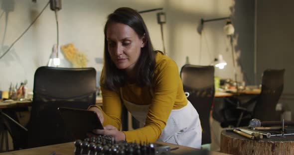 Caucasian female jeweller in workshop wearing apron, using tablet, smiling at camera alt