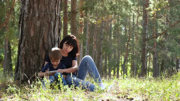Happy Family: Mother and Son Relax Sitting in the Park By a Tree. A Woman and a Child Look alt