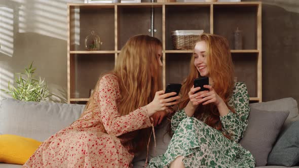 Two Young Twins Sisters Use Smartphones on Sofa in the Living Room alt