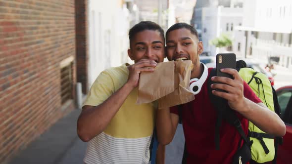 Two happy mixed race friends eating and taking selfie in the street alt