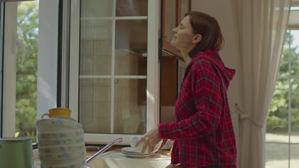 Young Adult Woman Washing Dishes and Enjoying Fresh Air By the Open Window alt