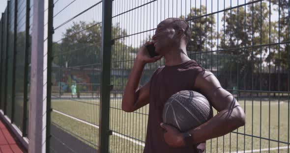 Positive African American Sportsman Talking on Mobile Phone Waiting for Friends with Ball at Outdoor alt