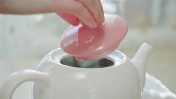 A woman's hand gracefully closes the lid of a white ceramic teapot with hot tea. Close-up. alt