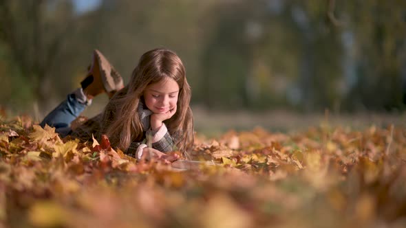 Sweet Girl in Polto Reads a Book Lying on Autumn Fallen Leaves. alt