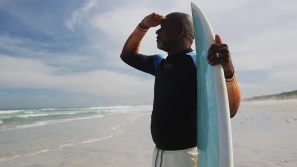 African american senior man standing on a beach holding surfboard and looking out to sea alt