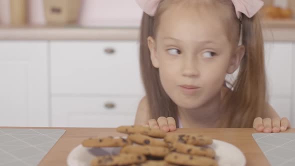 Cheerful Little Girl Peeking Out of Table and Taking Sweet Tasty Cookie From Plate, Portrait  alt