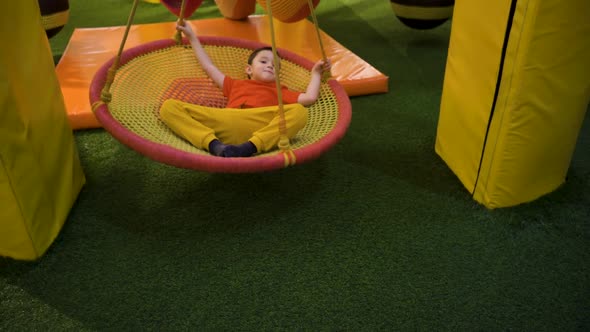 A Brunette Boy Swings on a Suspended Carousel Lying on His Back