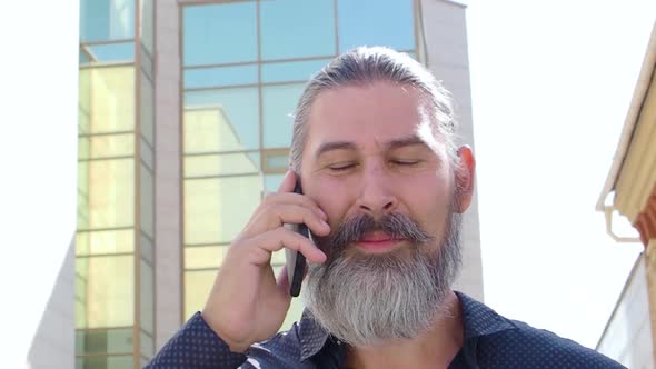 Middleaged Bearded Man Walks Down the Street Talking on His Cell Phone alt