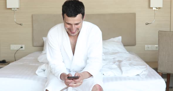 Smiling young attractive man wearing bathrobe using mobile phone. alt