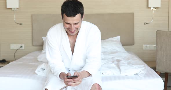 Smiling young attractive man wearing bathrobe using mobile phone.